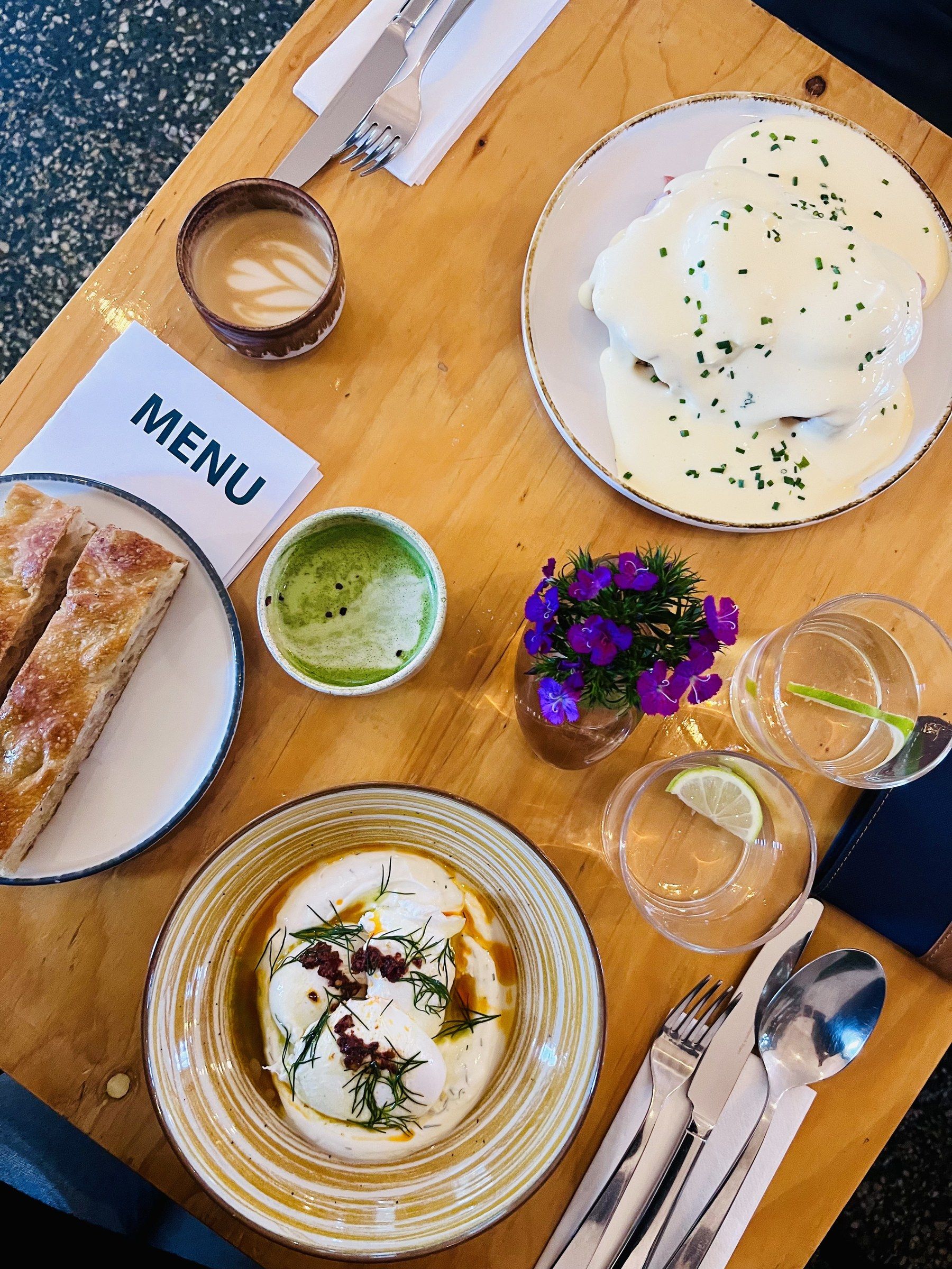 Pohled shora na dřevěný stůl s brunchem: vejce benedikt, matcha, croissant a menu.