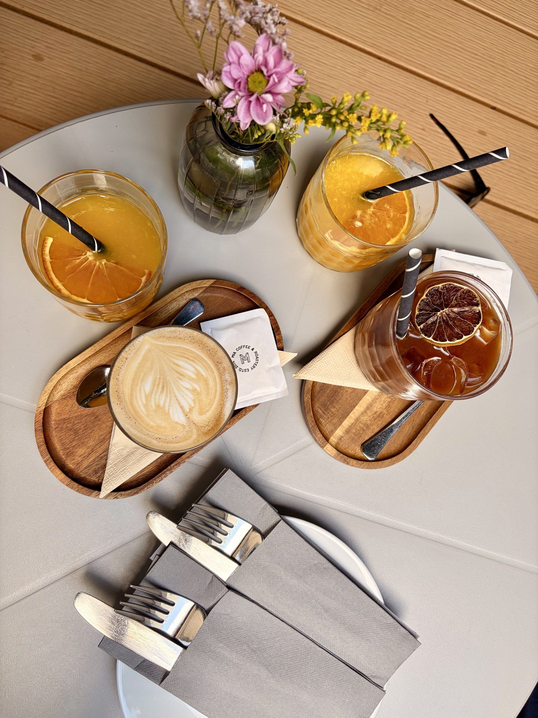 Pohled shora na kavárenský stůl: cappuccino, pomerančový džus, ledový čaj a kytička.